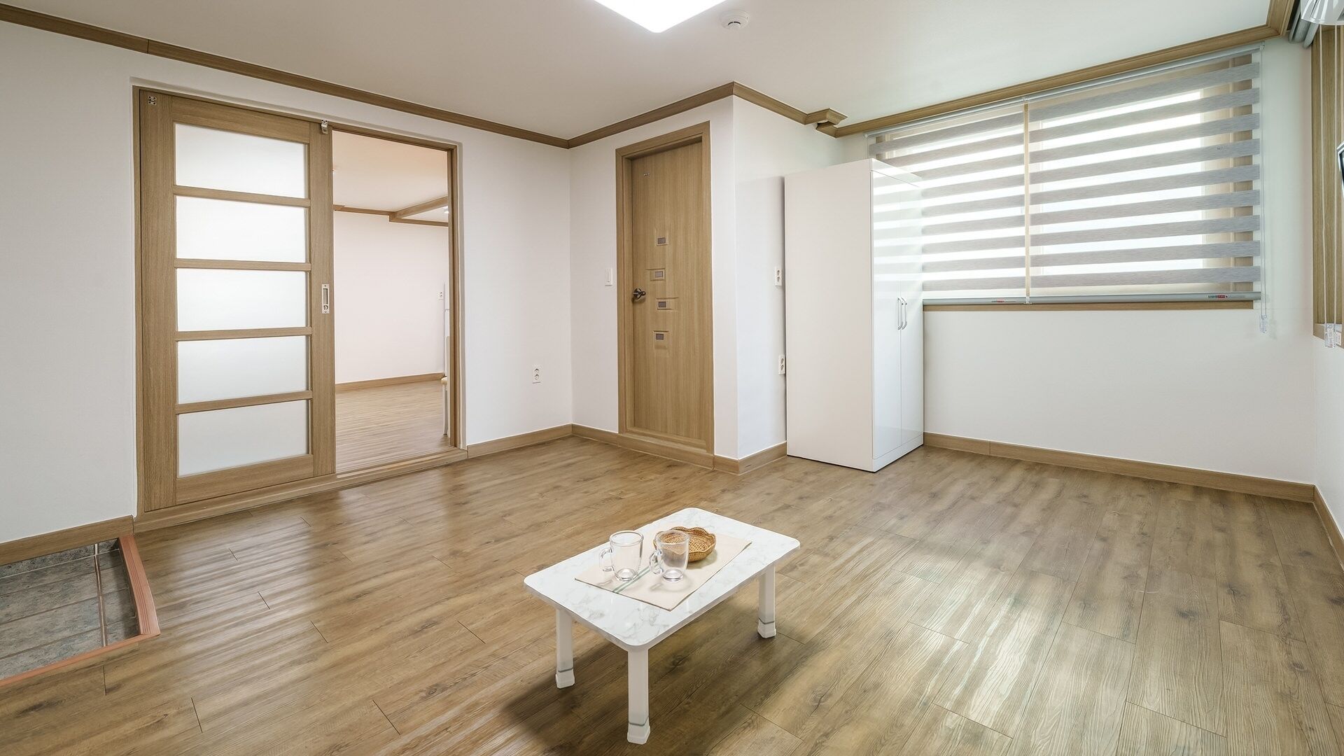 A room with wooden flooring, a white table with a basket, and a window with blinds.