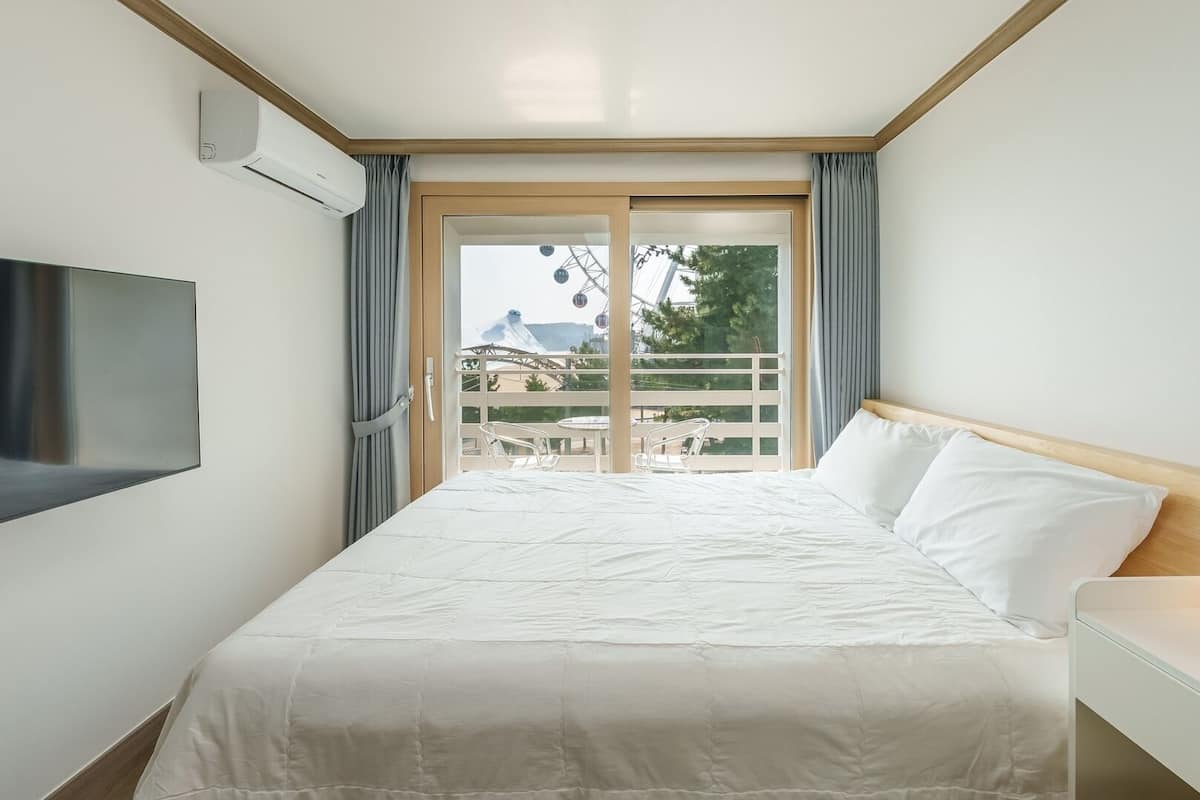 A hotel room with a bed, a TV, a bedside table with a lamp, and a view of a Ferris wheel and trees.