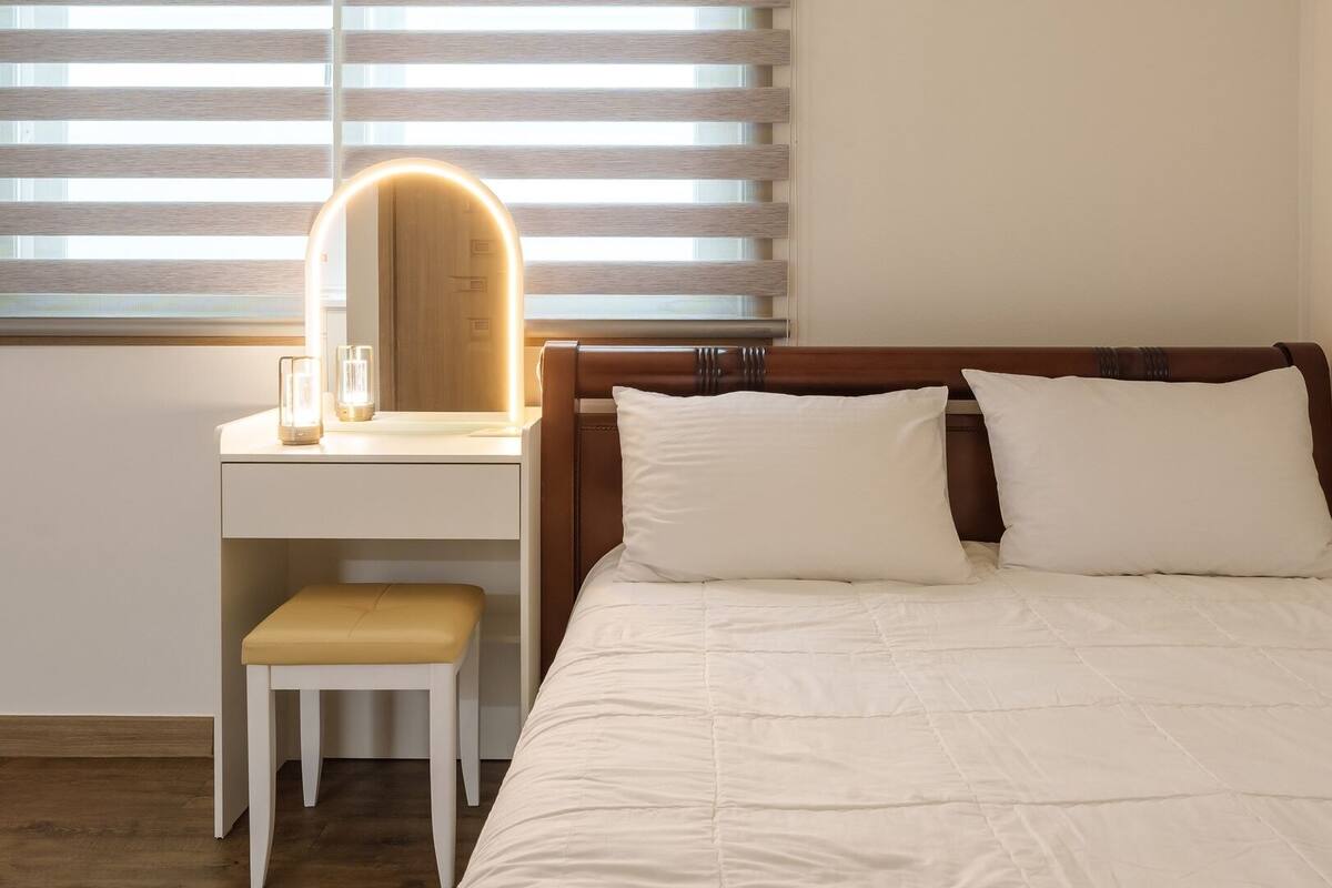 A bedroom with a bed, a bedside table with a lamp, a chair, and a window with blinds.