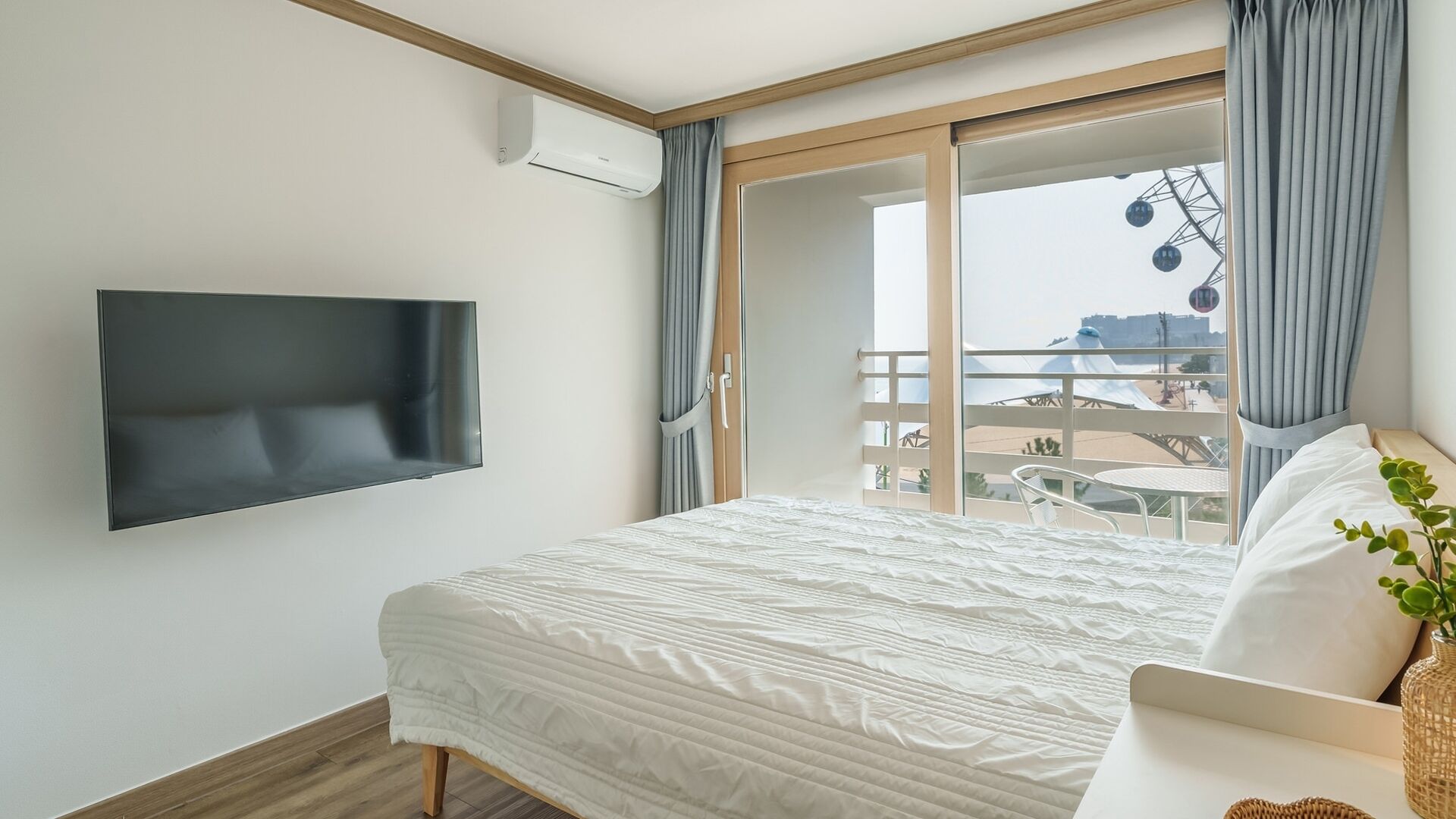 A hotel room with a bed, a flat-screen TV, a balcony with a table and chairs, and a view of the cityscape.