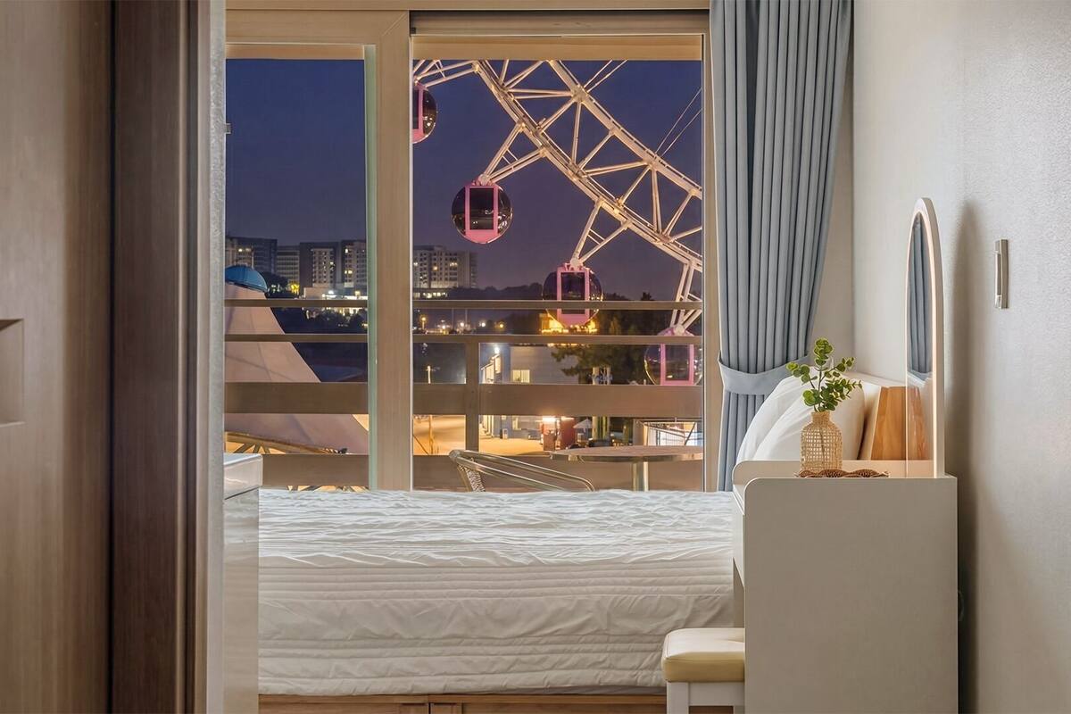 A hotel room with a bed, a desk, a chair, a window with a view of a Ferris wheel, and a curtain.