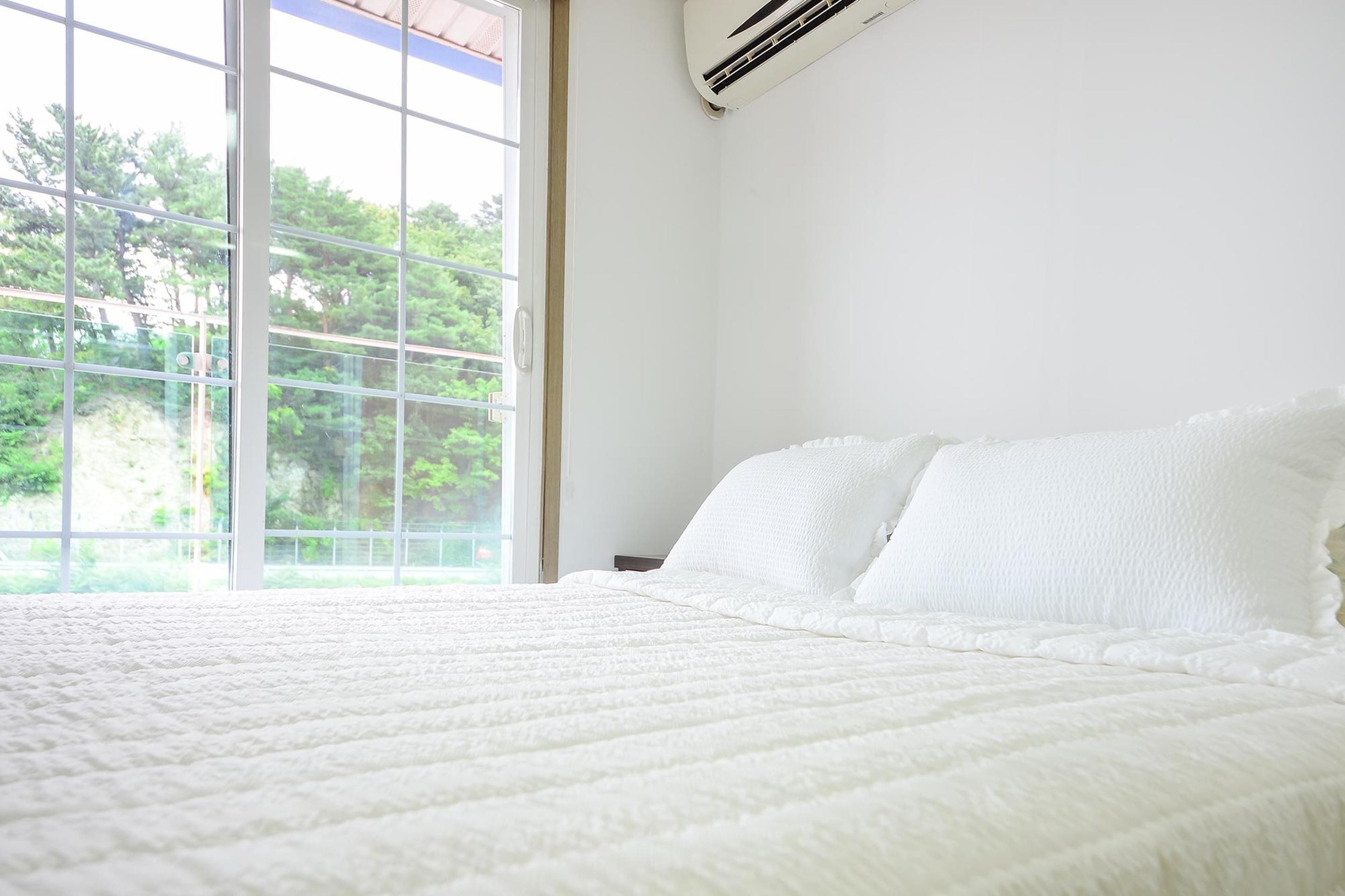 Un dormitorio con un ventanal grande, ropa de cama blanca y un equipo de aire acondicionado en la pared.