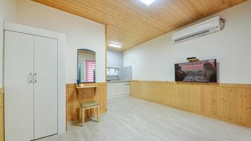 A room with wooden paneling, a white wardrobe, a bench, a television, and a kitchen area in the background.