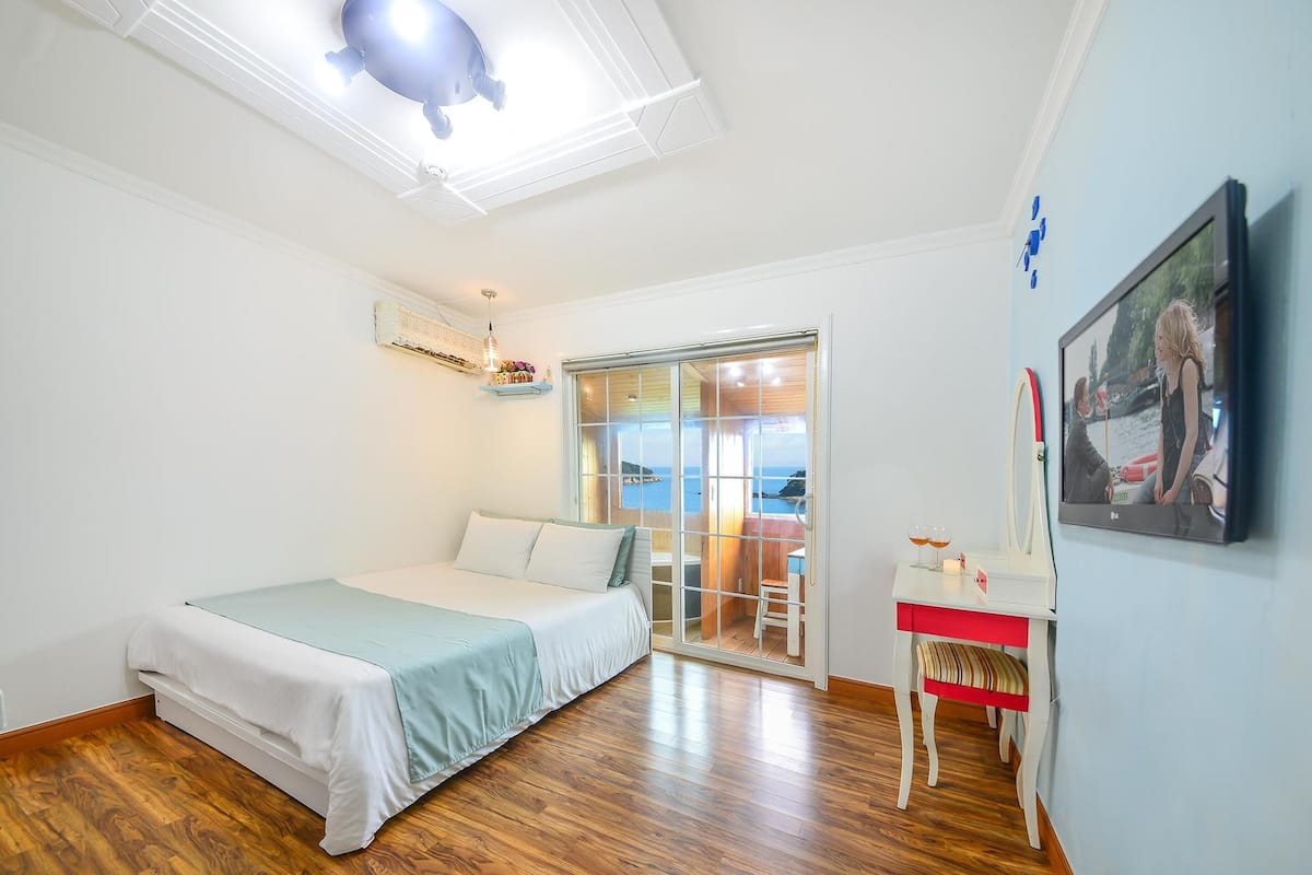 A bedroom with a bed, a desk, a chair, a ceiling light, a window, and a door.