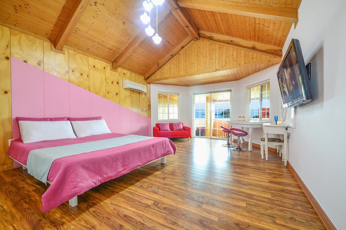 A bedroom with a large bed, a red sofa, a dining table with chairs, and a TV mounted on the wall.