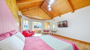 Un dormitorio con techo de madera, una cama con ropa de cama rosa y blanca, una silla roja, un escritorio con dos sillas y una ventana con vistas.