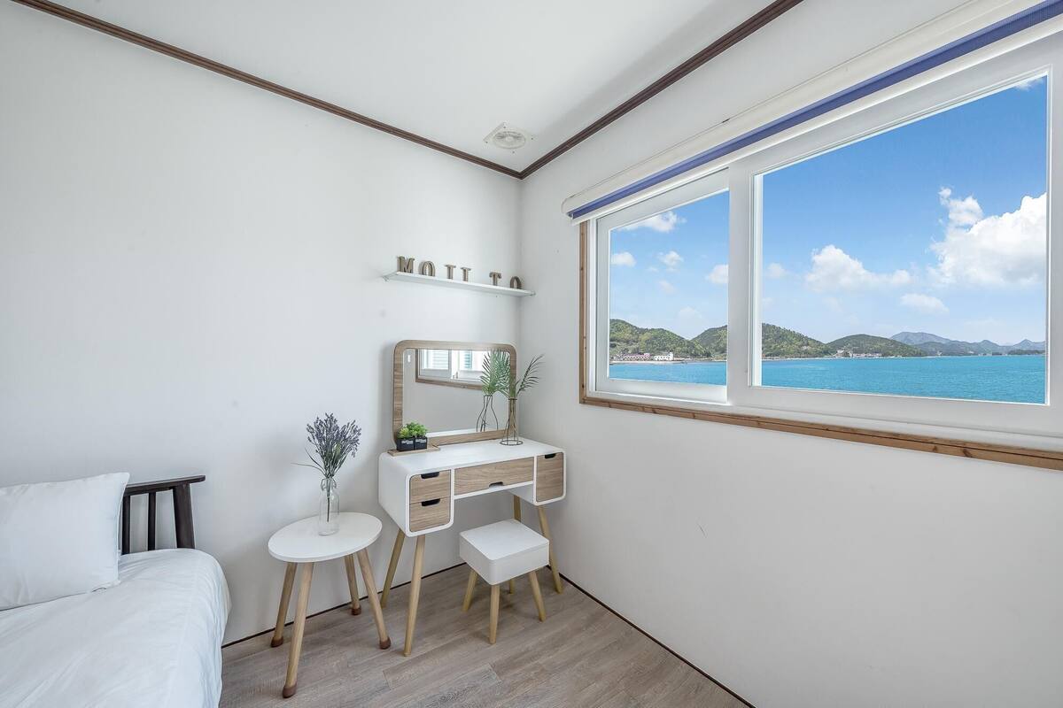 A bedroom with a bed, a desk with a mirror, a small table with a plant, and a window with a view of the sea and hills.