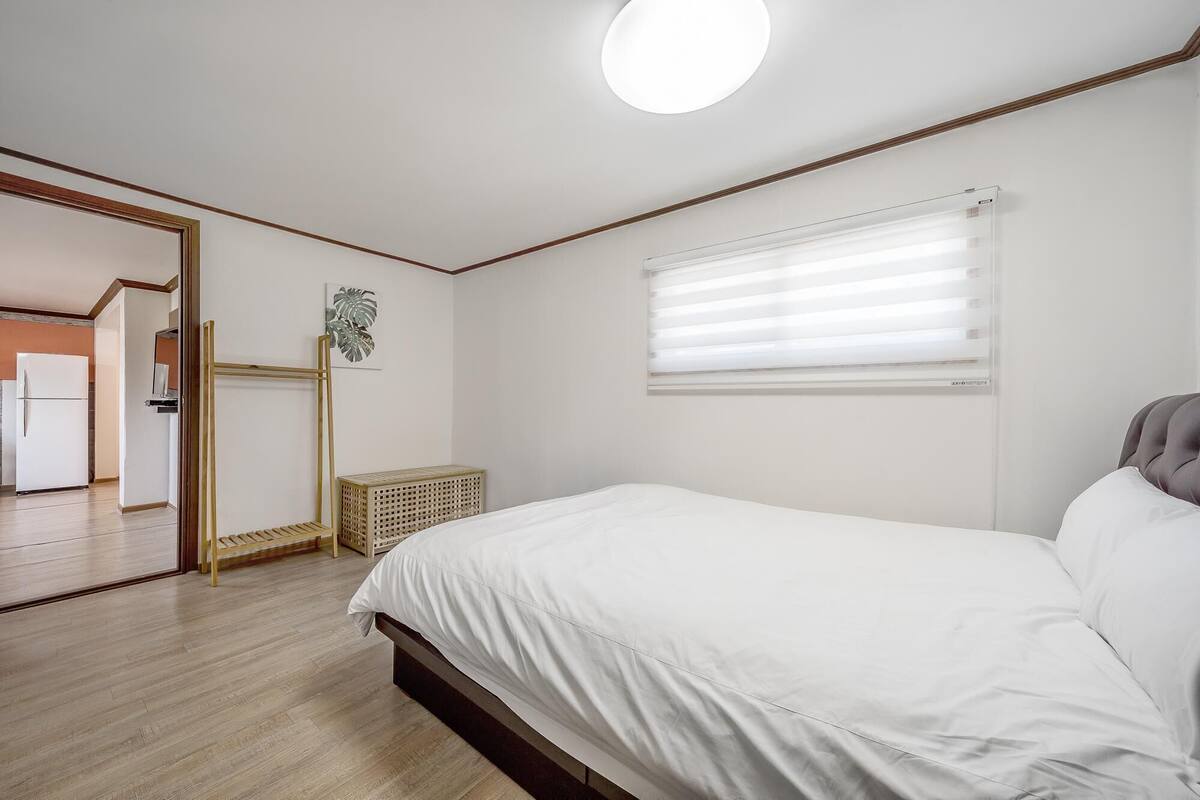 A bedroom with a bed, a window with blinds, a door leading to another room, and a wall-mounted artwork.