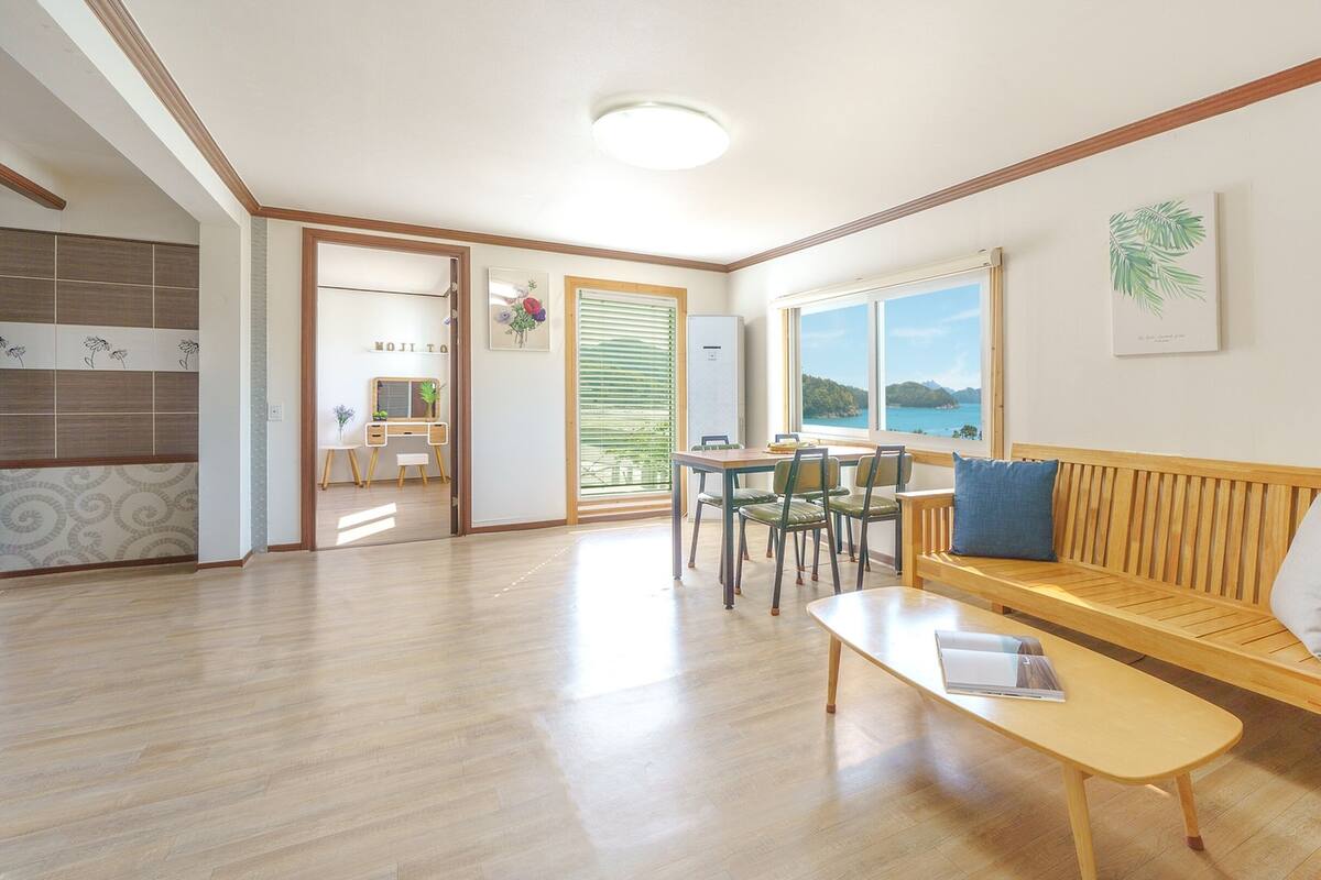 A spacious living area with a wooden bench, a dining table with chairs, a small desk, and a large window offering a view of the sea.