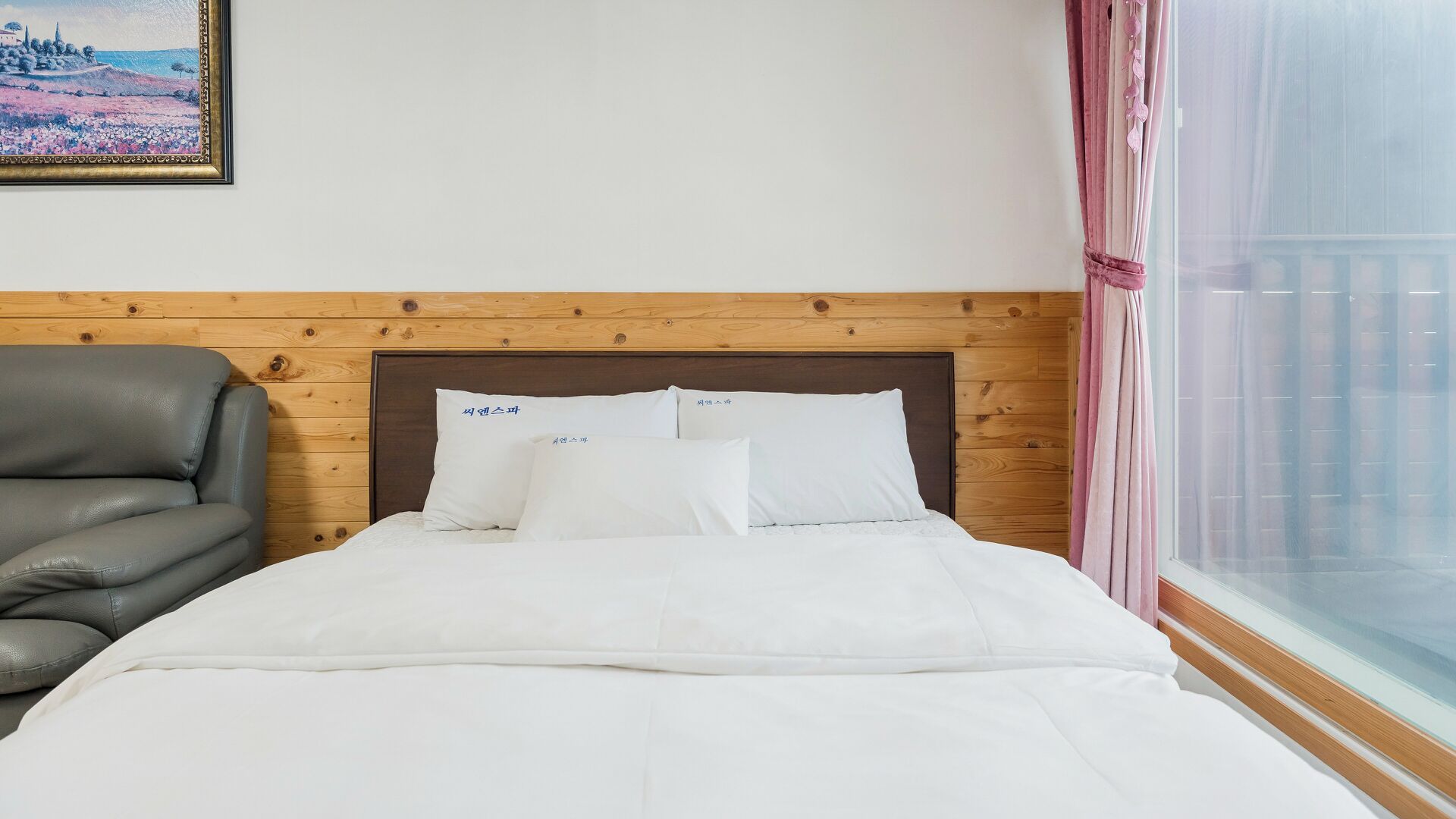 A hotel room with a wooden headboard, a grey leather sofa, a bed with white bedding, and a window with pink curtains.