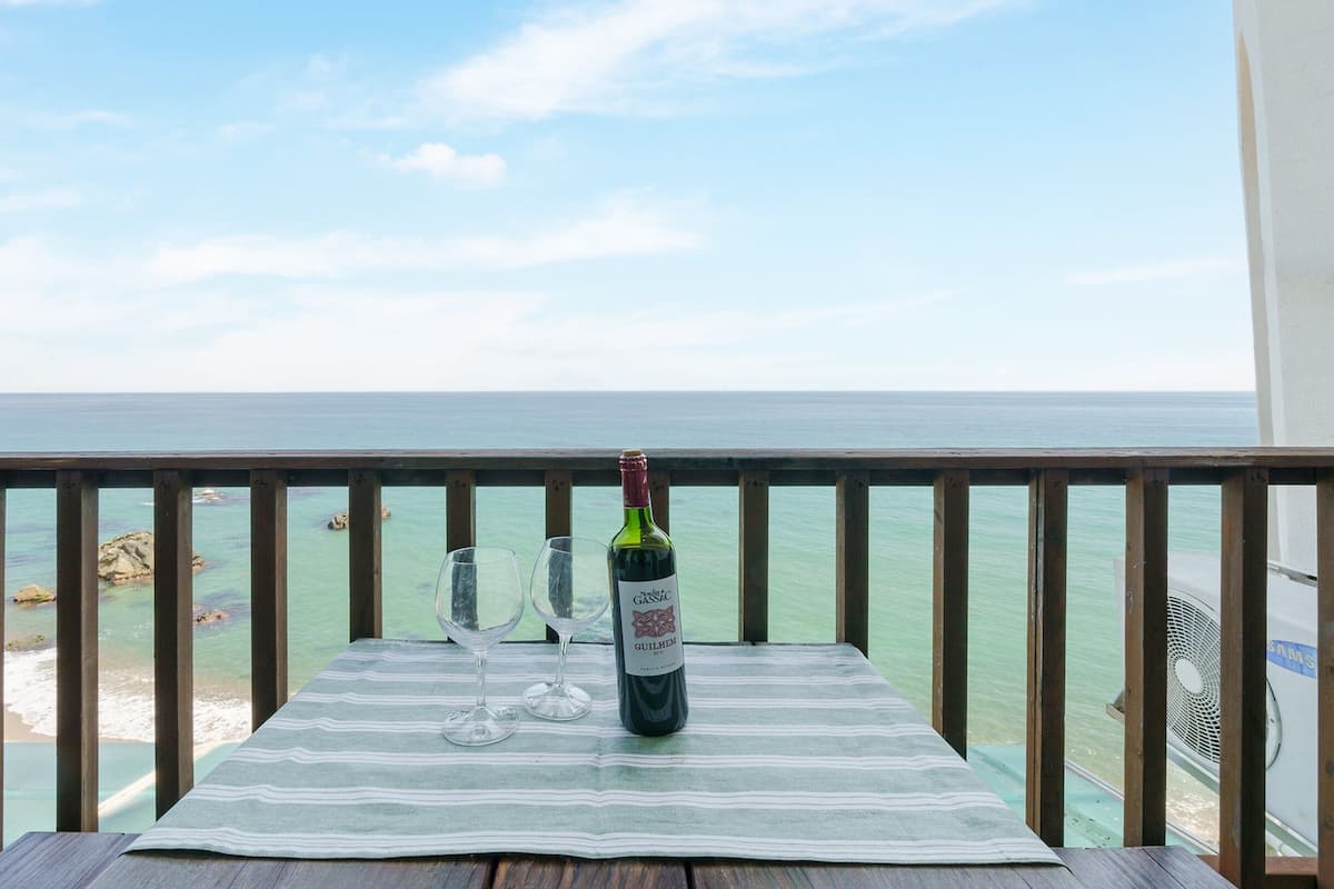 A table with a bottle of wine and two glasses on a striped tablecloth, overlooking a beach and the sea.