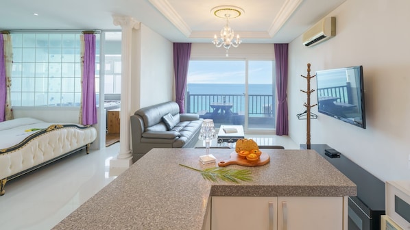 A modern hotel room with a bed, a sitting area with a sofa and a dining table, a kitchen area, and a balcony with a view of the sea.