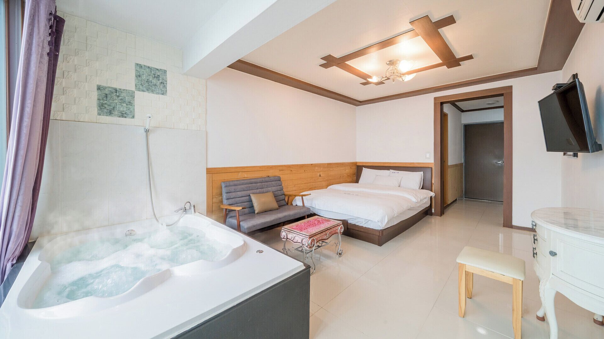 A hotel room with a large bathtub, a double bed, a small sofa, a bedside table, and a television.