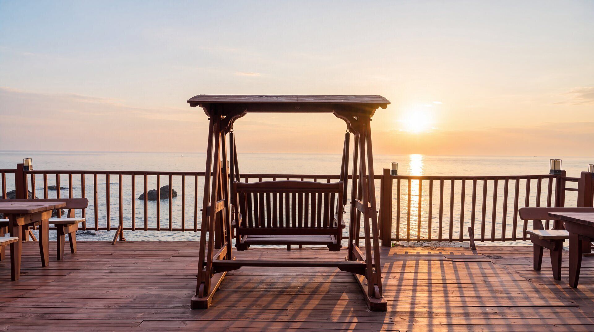 A wooden deck with a swing set and tables, overlooking a calm sea at sunset.