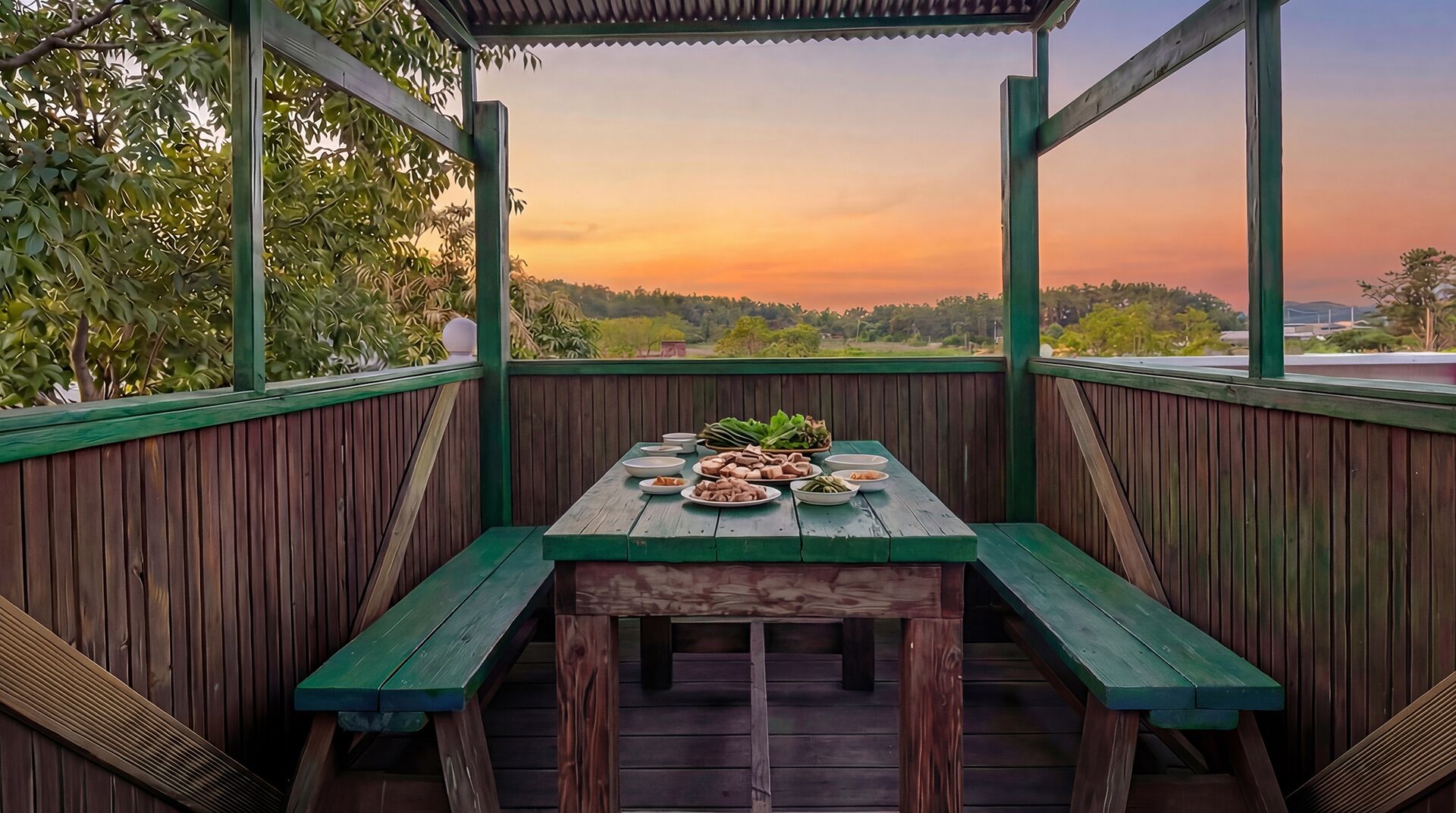 A wooden deck with a table set for a meal, surrounded by a wooden fence and a view of trees and a sunset.