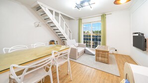 Un apartamento moderno con una mesa de comedor de madera, sillas blancas, un sofá, una escalera y un amplio ventanal con cortinas.