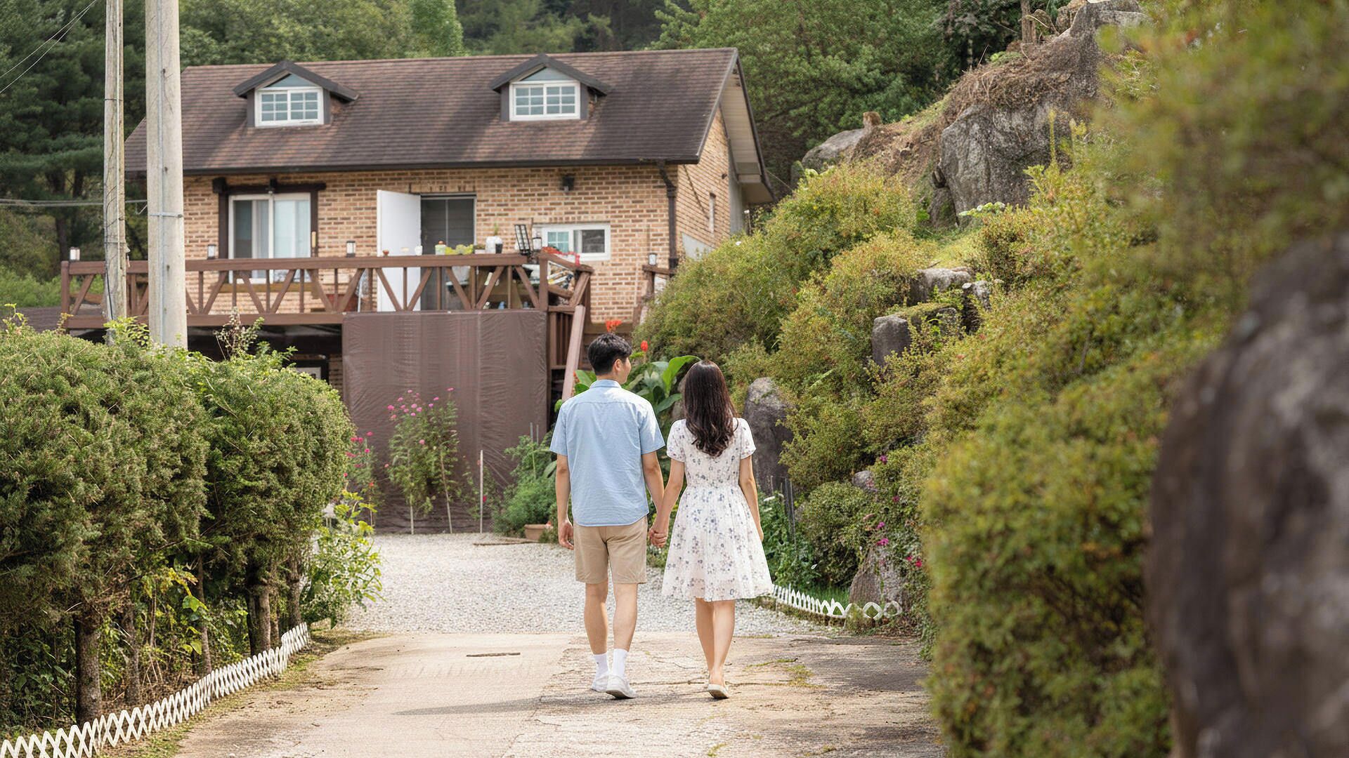 A couple walking on a gravel path towards a brick house with a wooden deck.