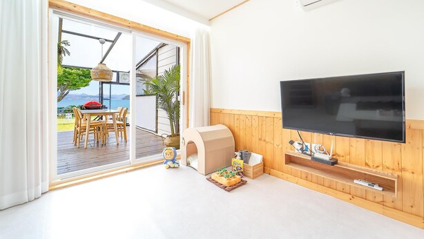 Una sala de estar con televisor, paneles de madera y un ventanal corredero que da a un espacio exterior con una mesa y sillas.