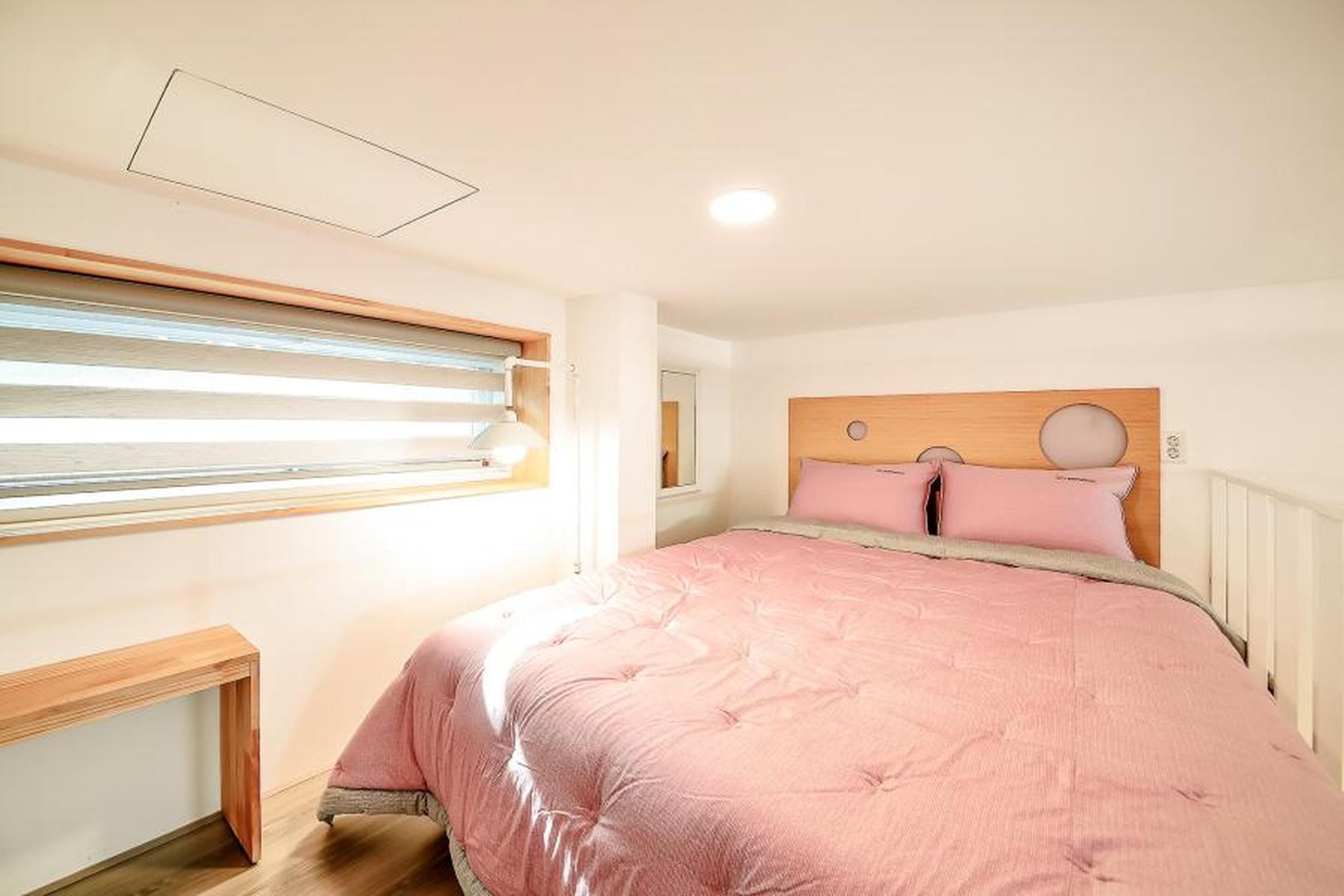 Un dormitorio con colcha rosa, cabecero de madera y ventana con persianas.