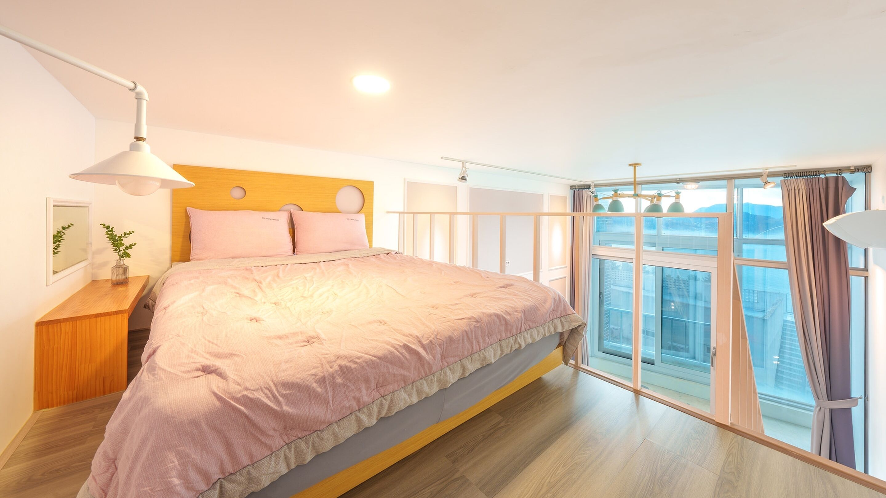 A bedroom with a large bed, a bedside table with a lamp, a window with curtains, and a view of the city.