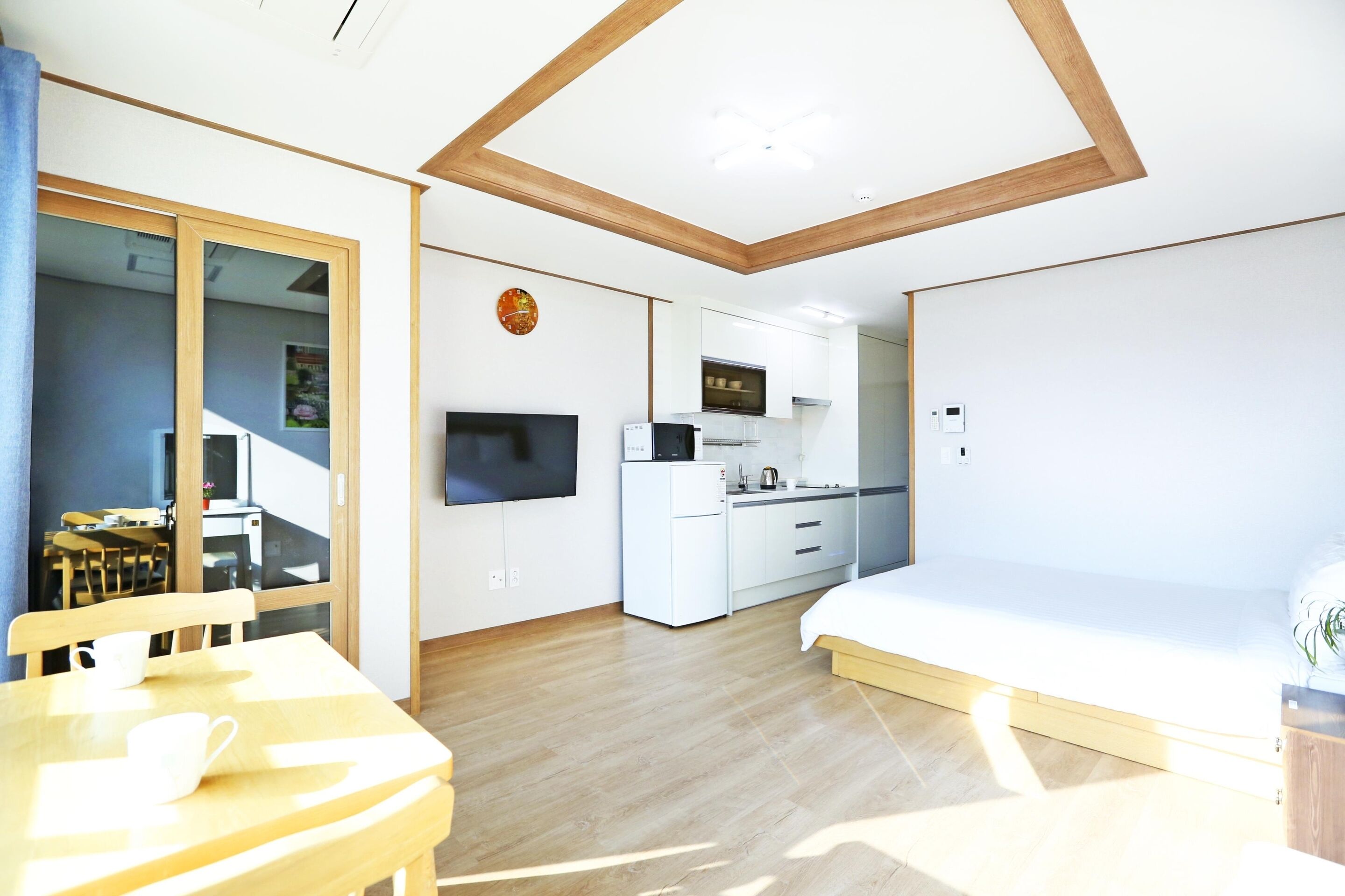 A compact room with a bed, a dining table, a TV, a kitchenette, and a sliding door leading to another room.