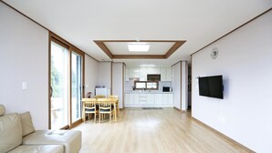 A modern living room with a sofa, a dining area with chairs, a kitchen with cabinets, and a flat-screen TV mounted on the wall.