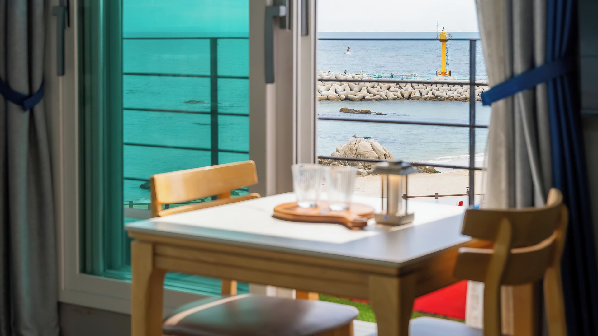 Una mesa con sillas y un juego de vasos, con vista a una costa rocosa y un faro.