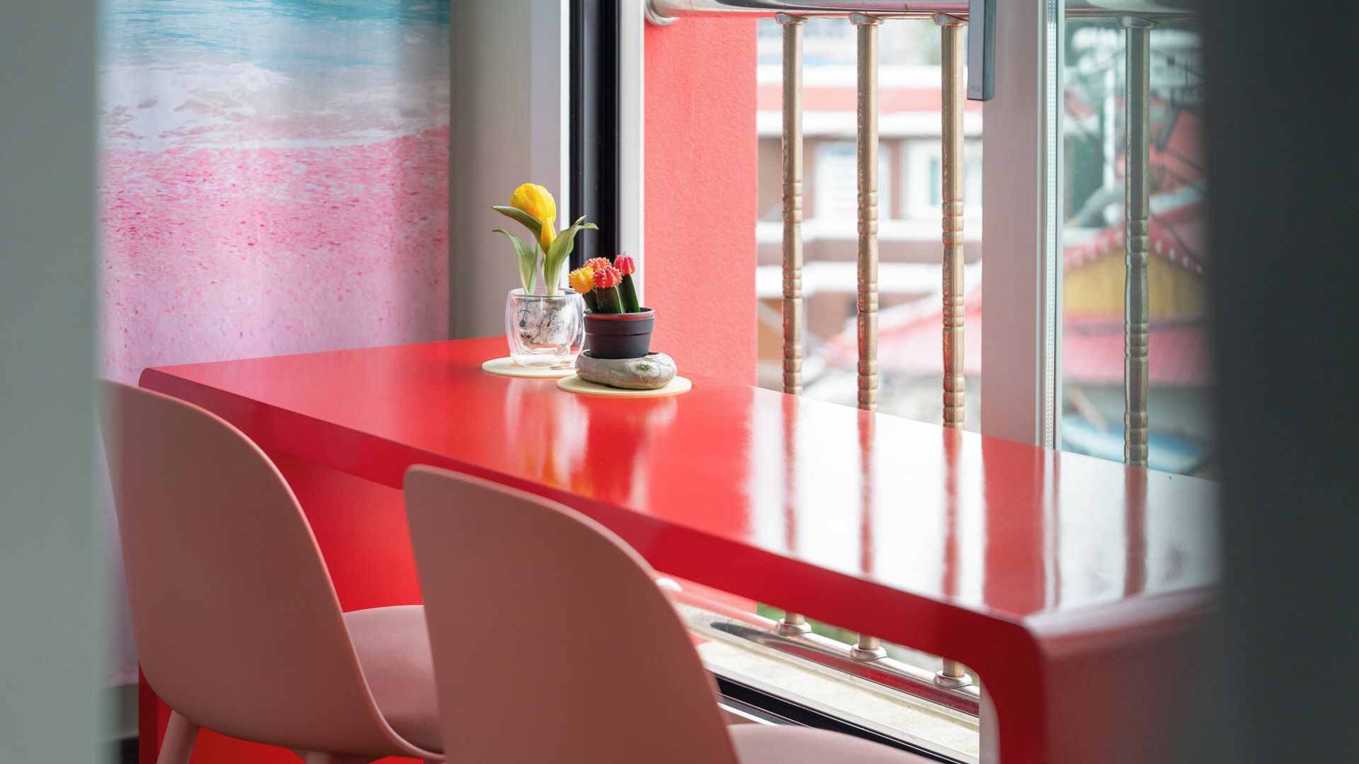 A red table with two chairs, a vase with flowers, and a window with a view of buildings.