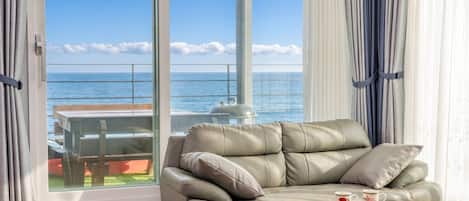 A room with a leather sofa, a table with two mugs, and a view of the sea through a large window.