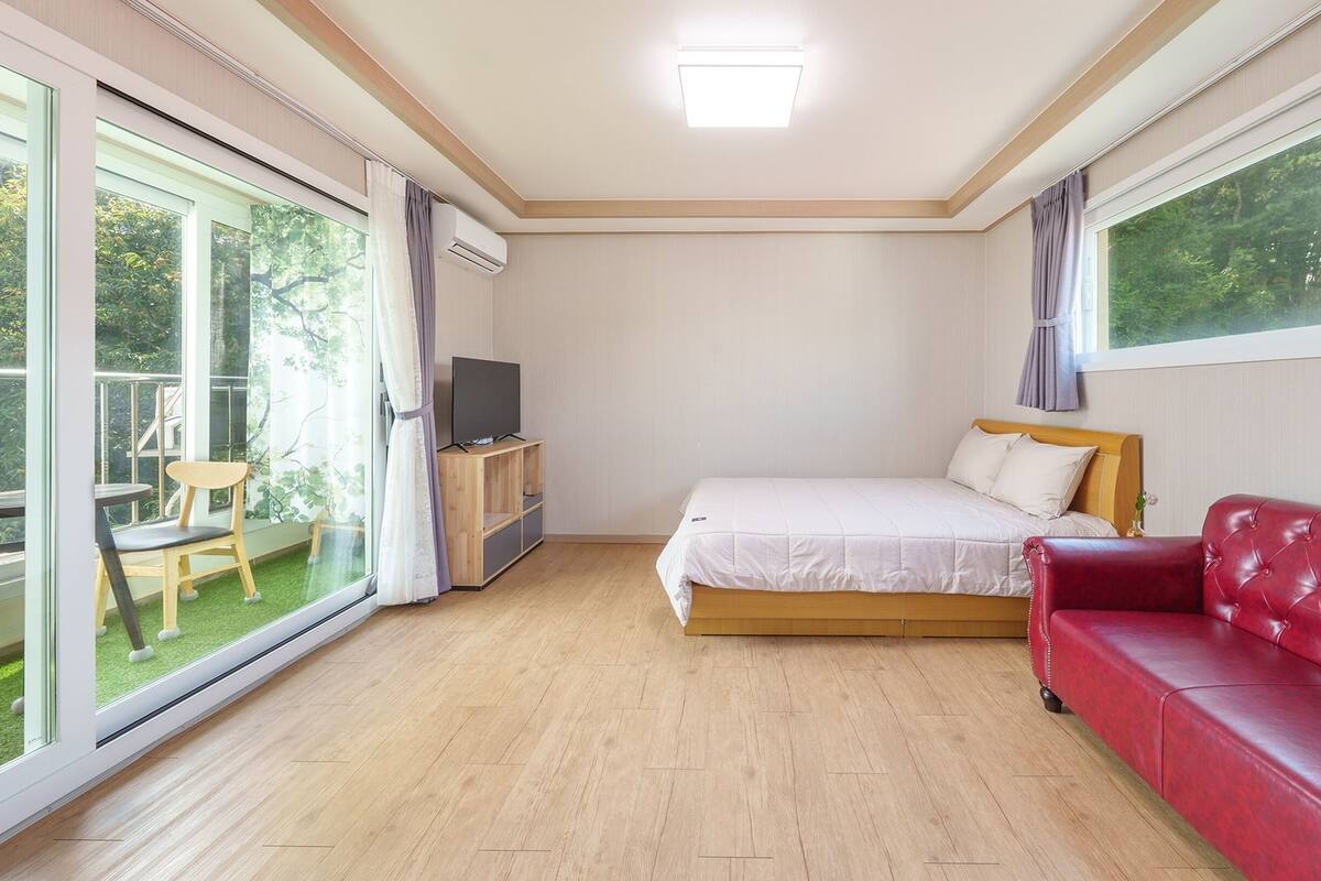 A bedroom with a bed, a red sofa, a TV on a wooden stand, and a balcony with a chair and table.