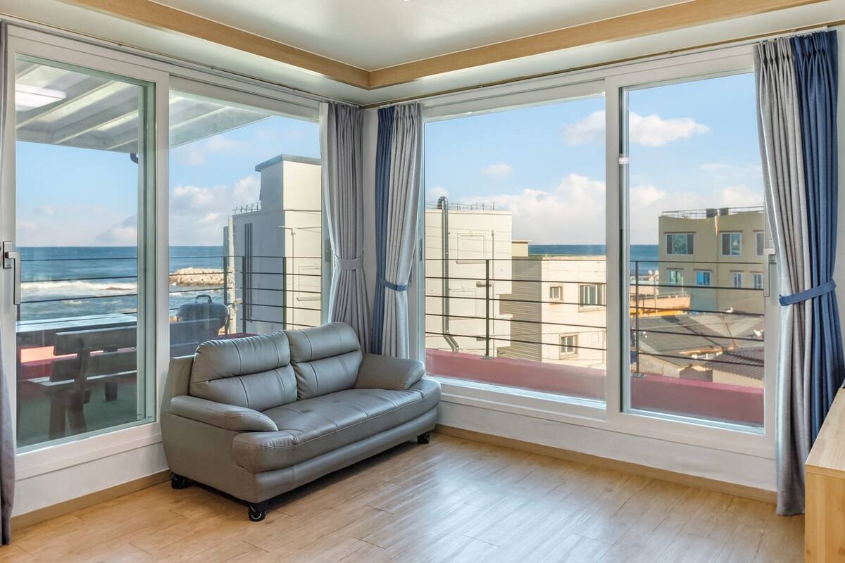 A modern living room with a grey sofa, large windows, and a view of the sea and buildings.