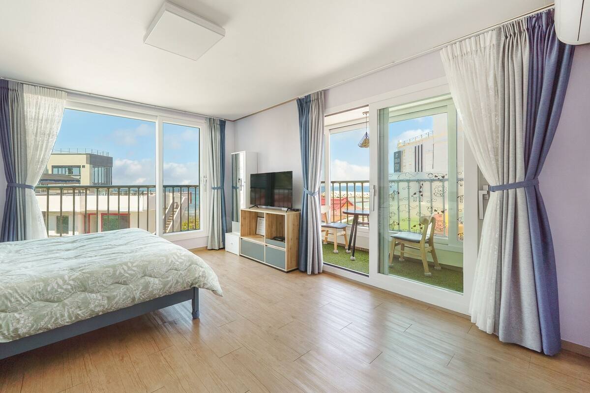 A modern hotel room with a large bed, a TV, and a balcony with a view of the beach.