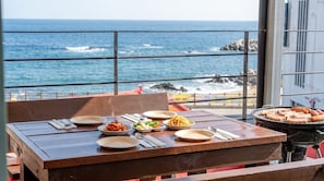 Un área de comedor al aire libre con vista al mar, que incluye una mesa de madera con platos de comida, una parrilla con salchichas y una barandilla metálica.