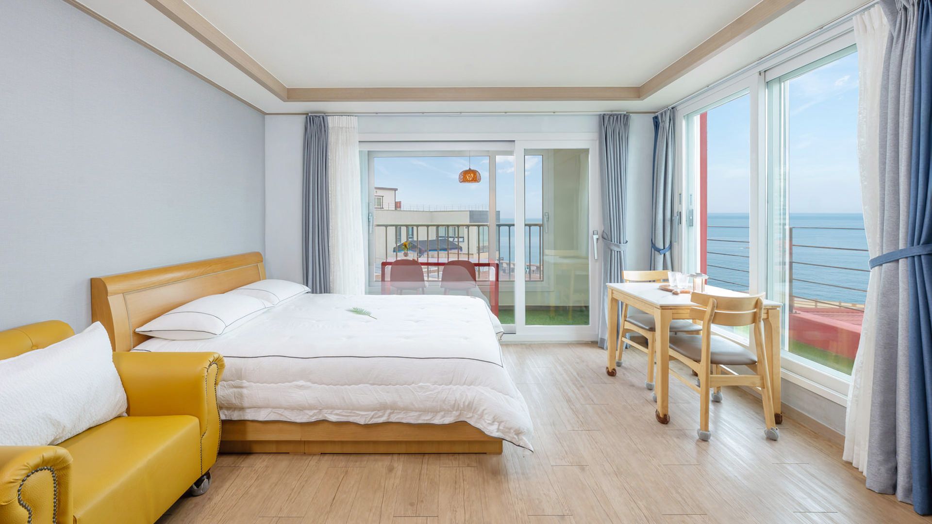 A hotel room with a bed, a yellow armchair, a dining table with chairs, and a balcony with a view of the sea.