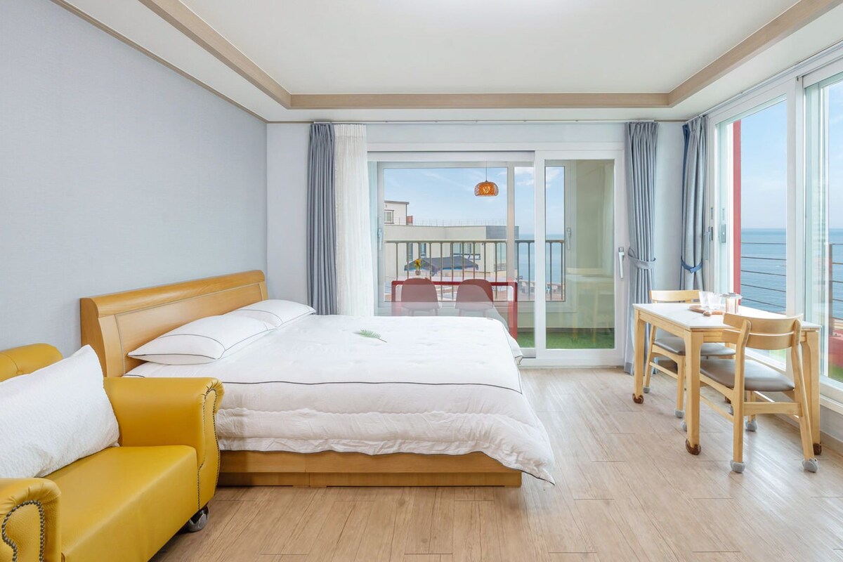 Een hotelkamer met een bed, een gele fauteuil, een eettafel met stoelen, en een balkon met uitzicht op zee.