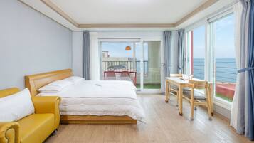 A hotel room with a bed, a yellow armchair, a dining table with chairs, and a balcony with a view of the sea.