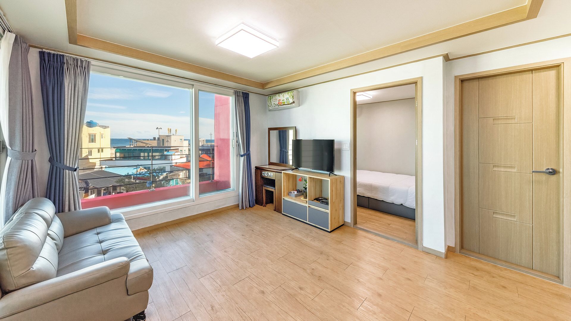 Une chambre d’hôtel avec un canapé, un téléviseur posé sur un meuble en bois, un lit et une vue sur les bâtiments à travers de grandes fenêtres.