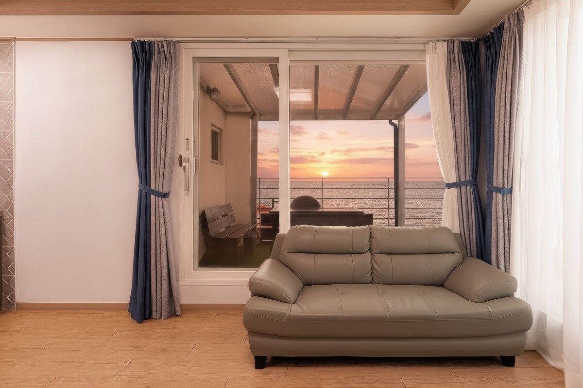 Een kamer met een bank, een schuifdeur naar een balkon met uitzicht op zee, en een betegelde vloer.