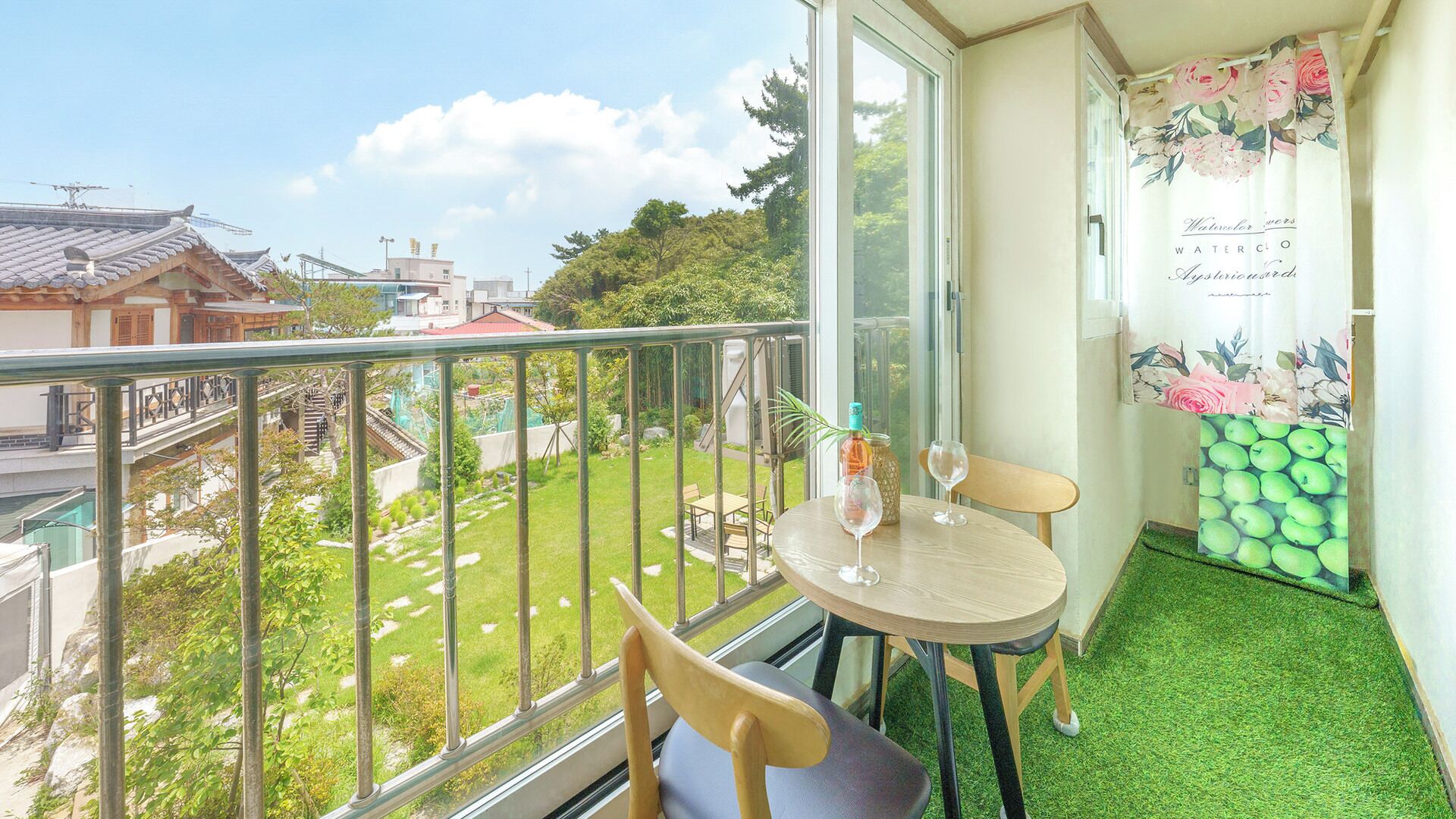 Ein Balkon mit Tisch und Stühlen, einer Weinflasche und einem Glas. Im Hintergrund sieht man einen Garten und Gebäude.