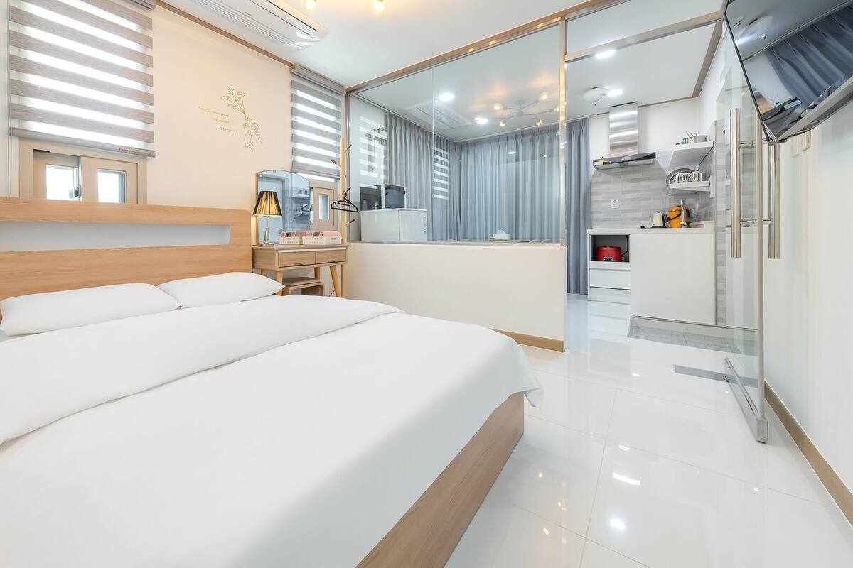 A modern hotel room with a large bed, a desk, a mirror, and a kitchenette.