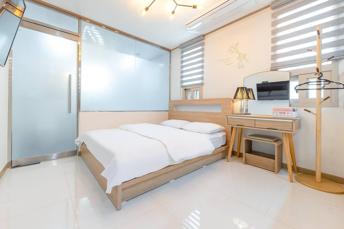 A modern hotel room with a bed, a desk, a TV, and a window with blinds.