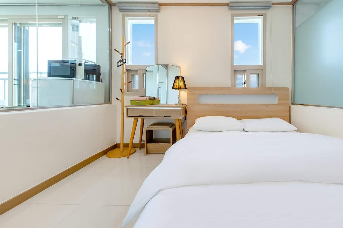 A hotel room with a bed, a desk, a chair, a lamp, and a window.