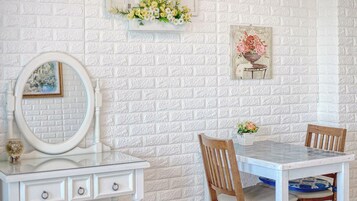 A room with a white brick wall, a white dresser with a mirror, a small table with chairs, and a framed floral painting.