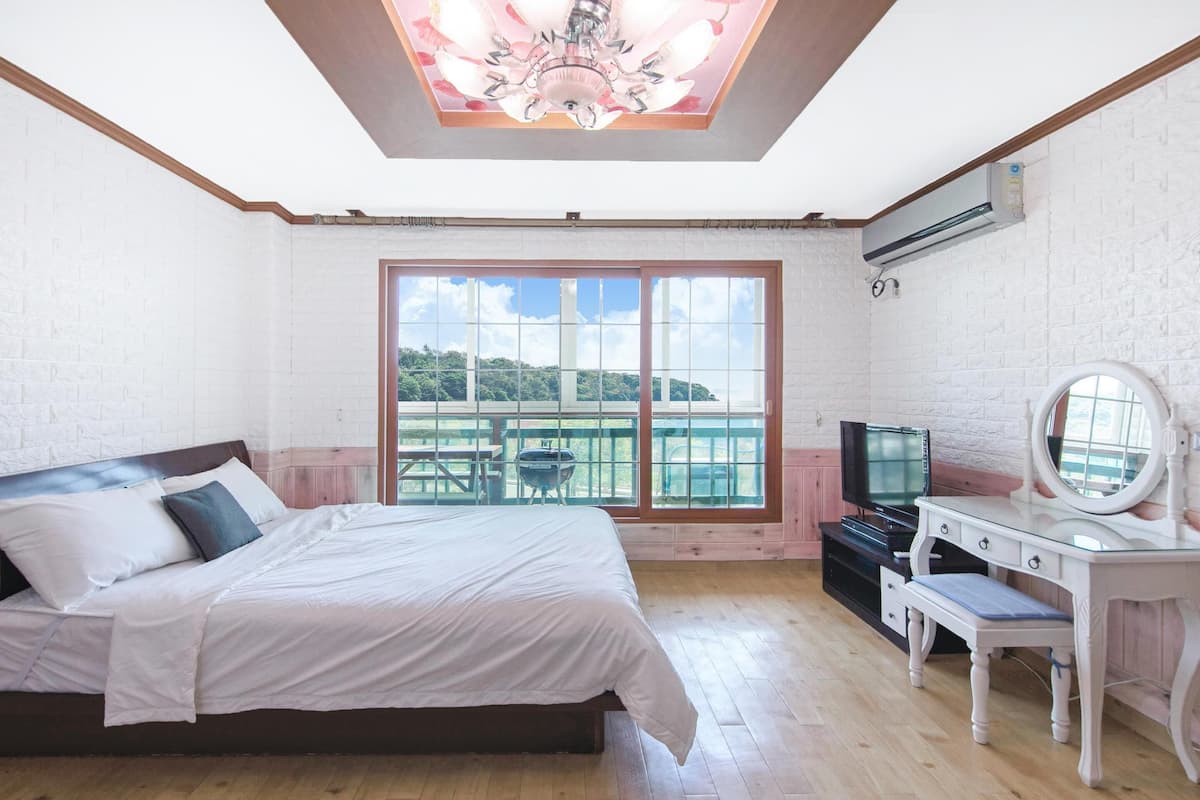 Ein Schlafzimmer mit Bett, Kommode, Fernseher und einem Balkon mit Blick auf Grünflächen.