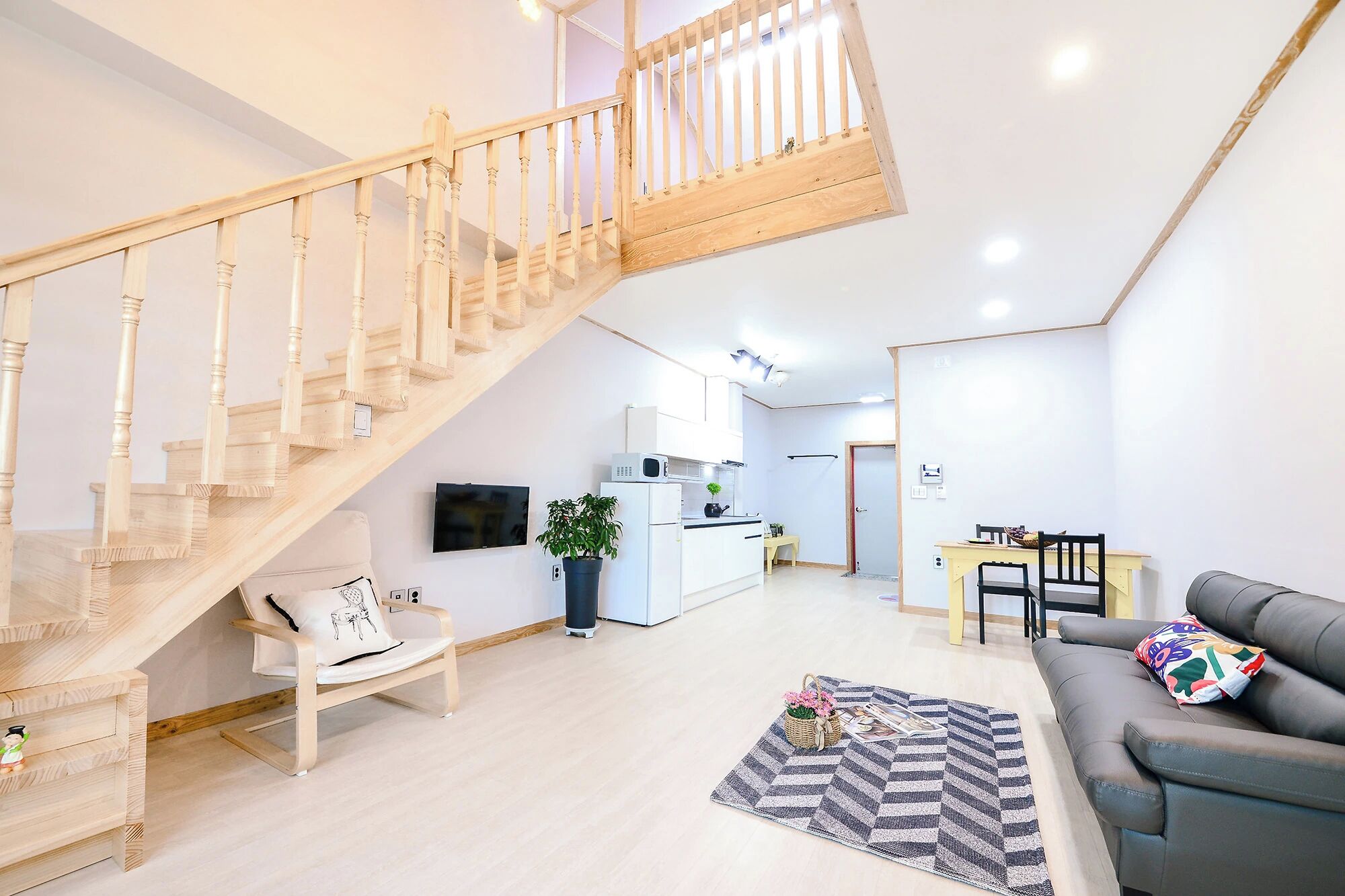 A modern living space with a wooden staircase, a kitchen area, a dining table with chairs, and a grey sofa.