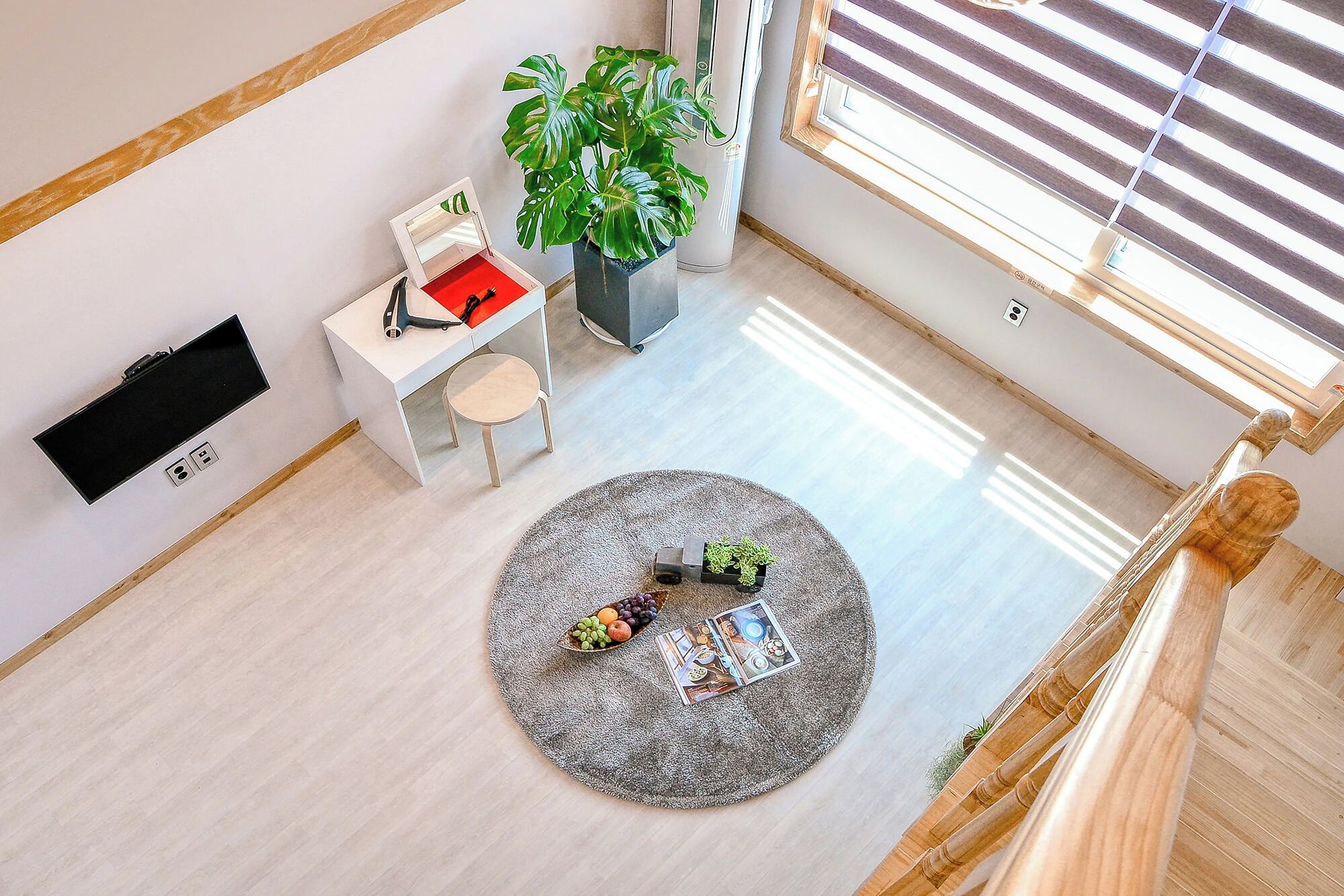 A room with a round rug, a small table with a plant, a white desk with a red cushion, and a large potted plant.