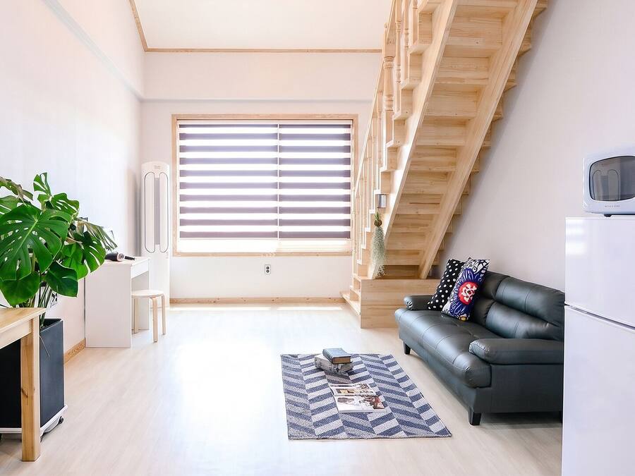 Un apartamento moderno con cocina, zona de comedor, sofá de cuero negro, escalera y un amplio ventanal con persianas.