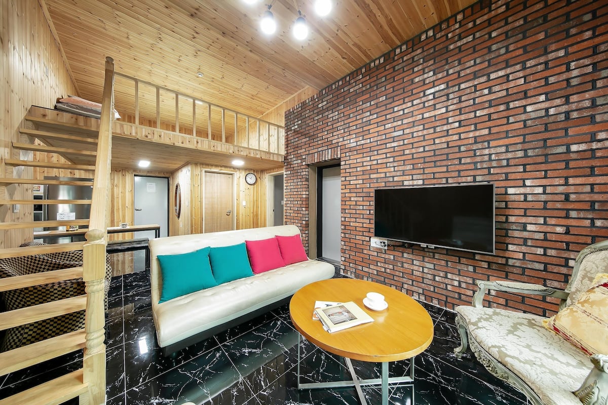 A modern interior with a wooden staircase, a sofa with colorful cushions, a round wooden table, and a brick wall with a flat-screen TV.