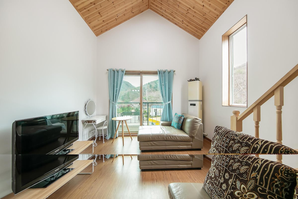 A modern living room with a flat-screen TV, a sofa, a patterned armchair, a small table, and a window with curtains.