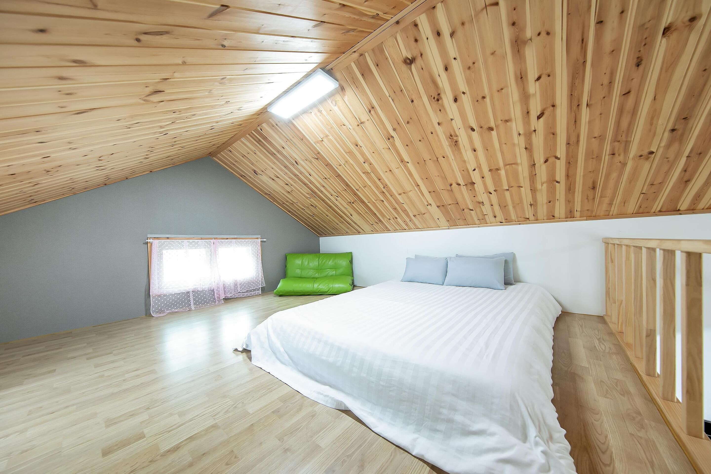 Une chambre avec un plafond en pente, un lit avec une literie blanche, un fauteuil vert et une fenêtre ornée de rideaux légers.